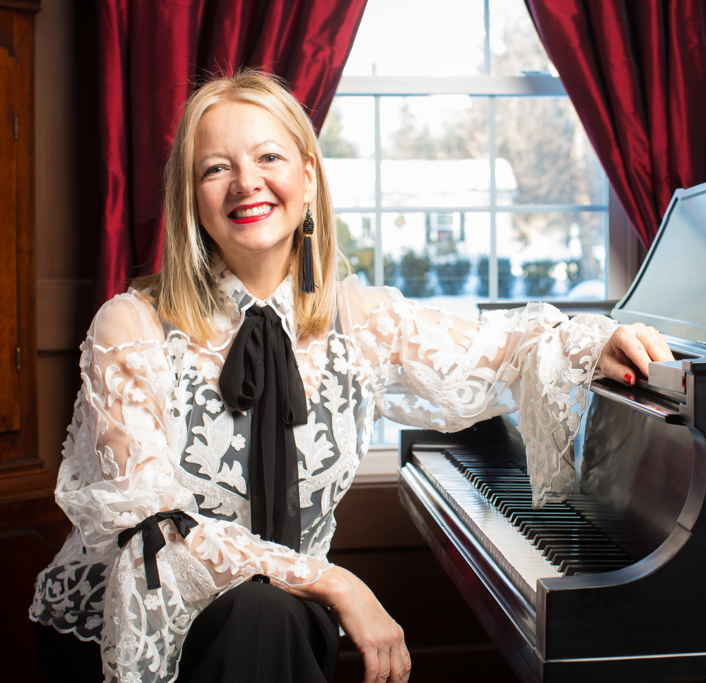 Kiki Cubb seated at a grand piano in an elegant white lace blouse with black bow